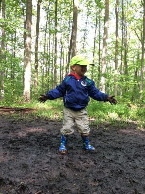 Child standing joyfully with mud-covered hands in a forest clearing.
