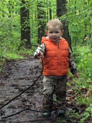 Child in orange vest exploring a muddy trail holding a stick.