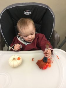 Toddler sitting in a high chair painting a pumpkin with orange and red paint.
