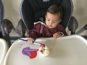 Toddler in high chair painting a white pumpkin with purple and pink brush strokes.