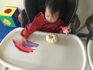 Toddler using hands to paint near a white pumpkin on a high chair tray.