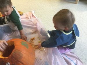 Two toddlers scooping out pumpkin seeds from a large carved pumpkin on a plastic mat.