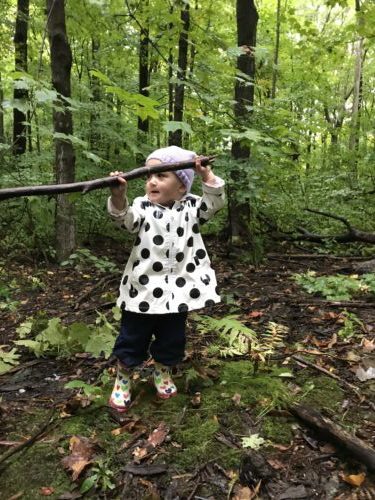 Young girl in white polka dot coat raises a large stick in green wooded area.