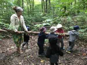 Children and educator collaborating to carry a large fallen branch during forest exploration.