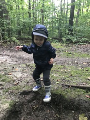 A child in a navy jacket and boots steps into a small gap in the forest floor during outdoor play.