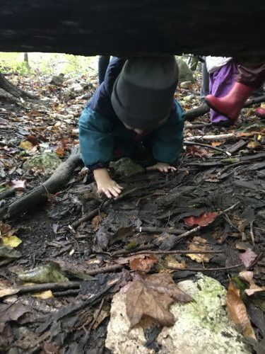 A young child crouches and crawls beneath a large log, engaging in active and messy forest play.