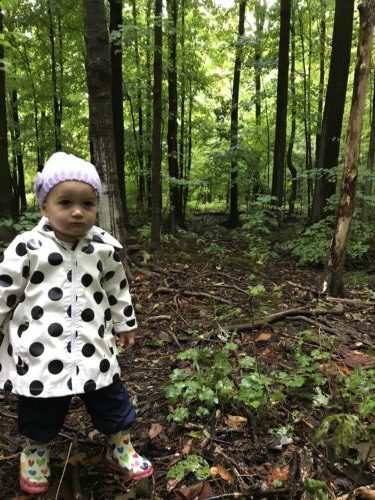 A toddler dressed in a white coat with black polka dots stands among trees on a damp forest trail.
