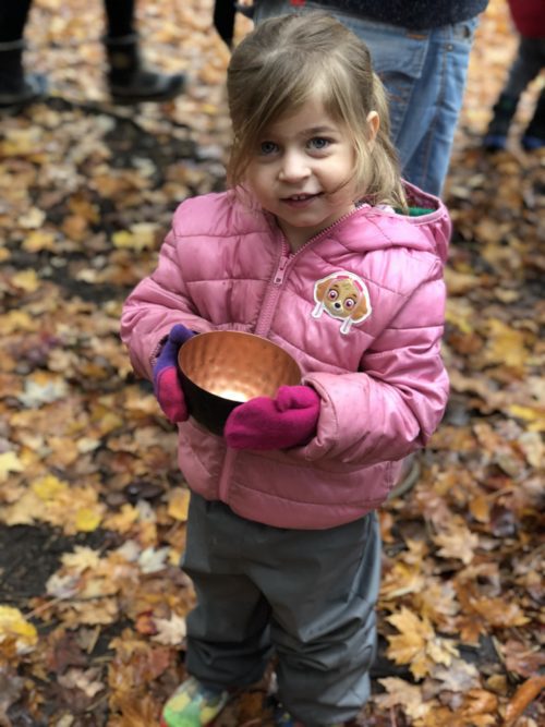 Child in pink jacket holding a brass bowl during fall ceremony in the forest.