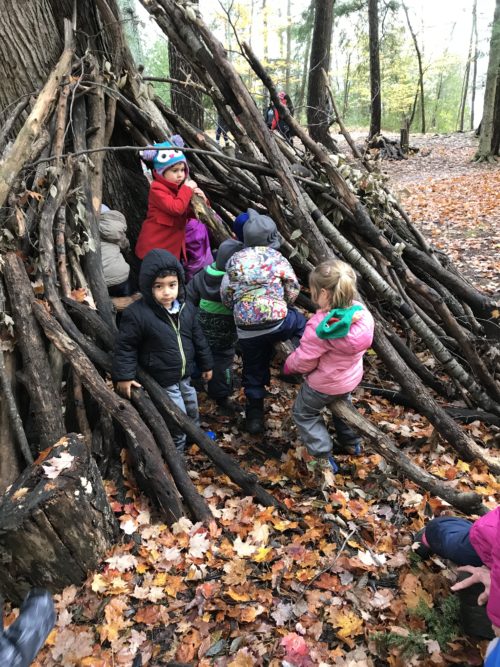 Group of children gathered around a large shelter made of sticks in a wooded area.