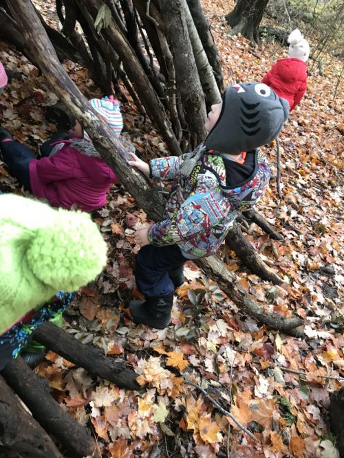 Young child standing at the entrance of a stick structure, surrounded by autumn leaves.