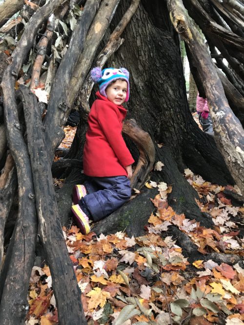 Child kneeling at the base of a tree inside a structure made of logs and branches.