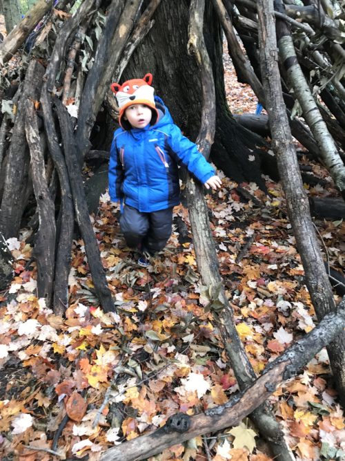 Young child in a blue coat and fox hat standing at the entrance of a forest stick shelter.