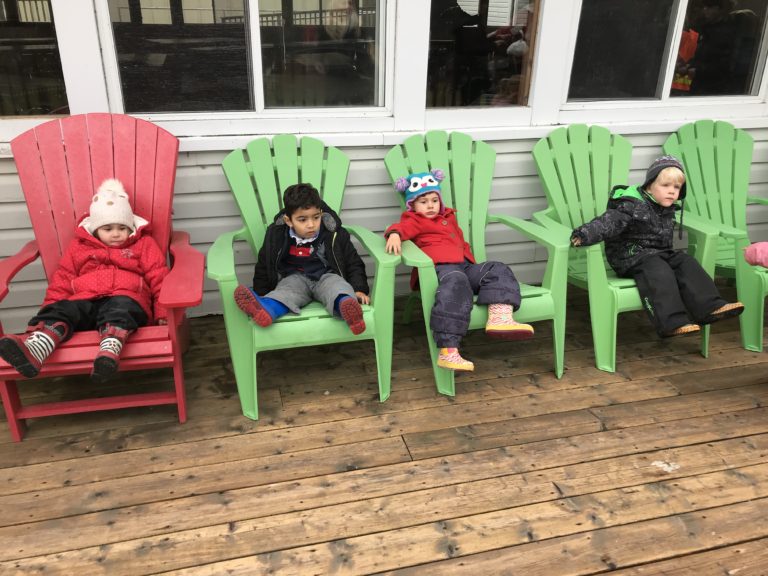 Four children bundled in winter gear relaxing on colourful Muskoka chairs on a wooden deck.