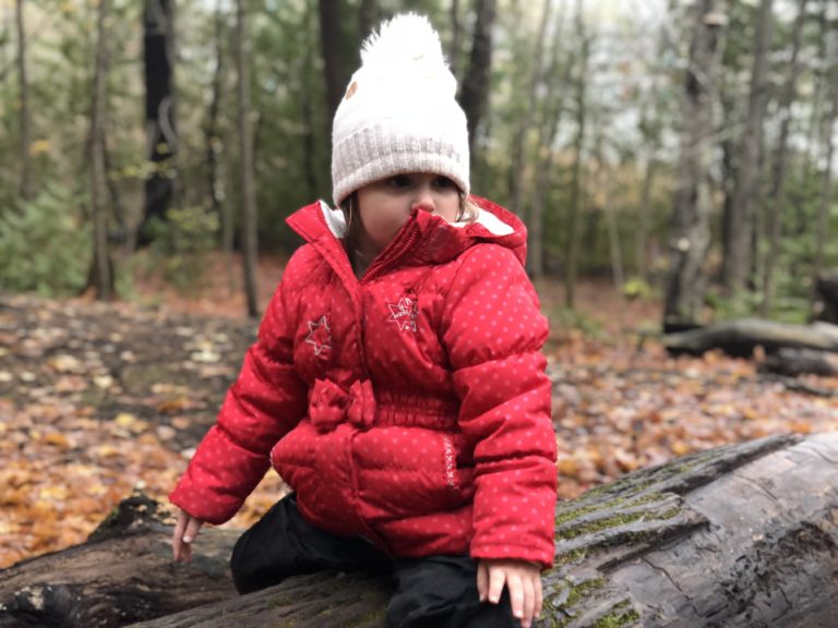 Child in a red jacket and white toque sitting on a log, observing the forest surroundings.