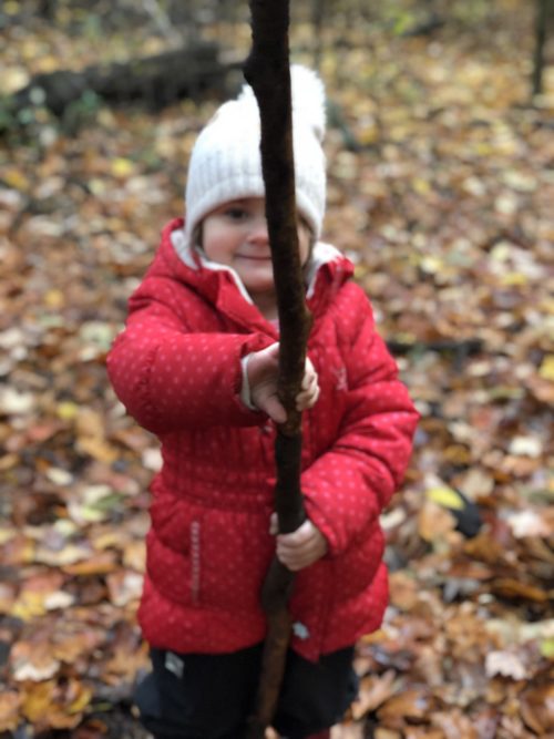 Child in a red jacket holding a large stick while standing among fallen autumn leaves.