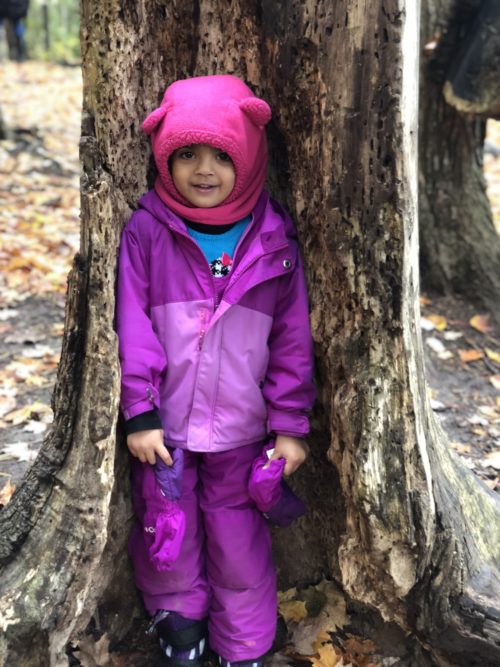 Smiling preschooler in a pink hat standing between a split tree trunk.