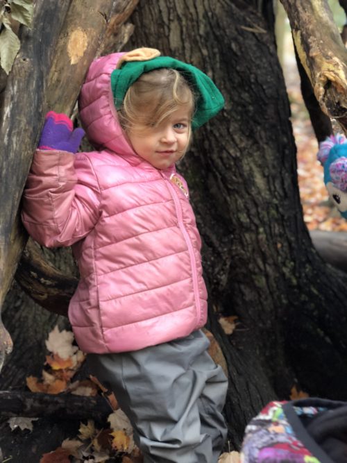 Girl in a pink jacket leaning inside a forest shelter built from branches.