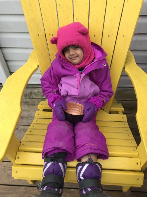 Child in purple and pink snowsuit sitting on a yellow chair, holding a copper bowl.