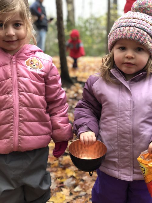 Two young girls in pink jackets holding brass bowl in a fall forest.