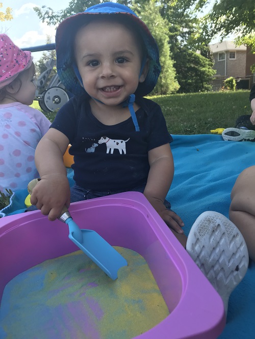 Smiling toddler scooping colourful sand from a pink bin outdoors.