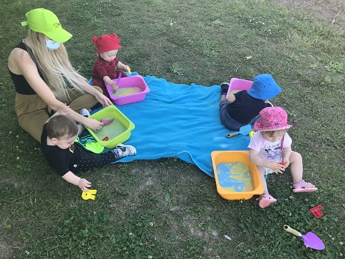 4 toddlers and educator sitting on blue blanket on grass, with scoops and coloured sand in bins.