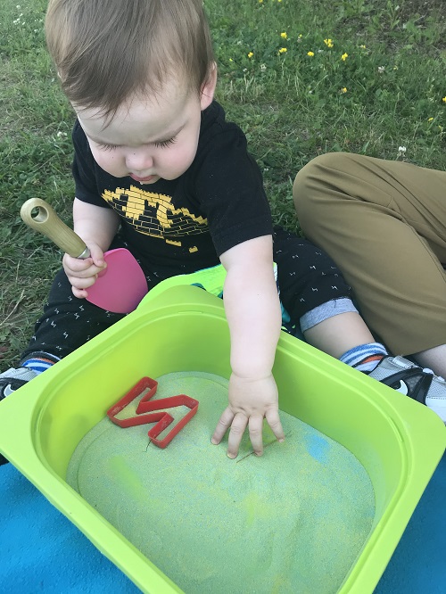 Toddler wearing black tshirt reaching into a green bin with sand.