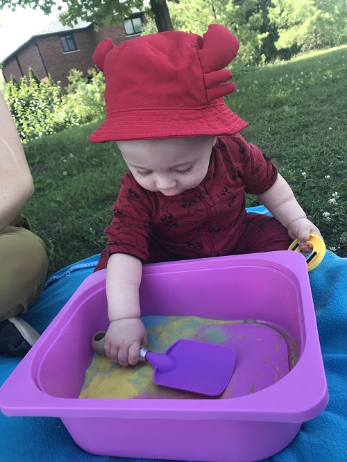 Infant scooping bright coloured sand from a pink bin.