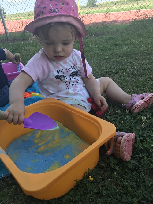 Infant in pink sunhat using purple scoop to play with coloured sand in an orange bin.