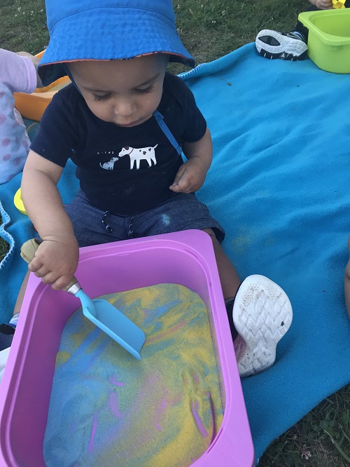 Toddler intently scooping coloured sand from a pink bin on blue blanket outdoors.