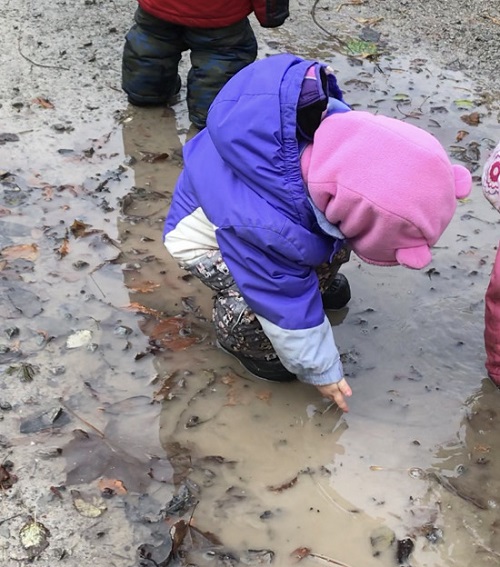 child in a pink hat and purple jacket touhing a puddle