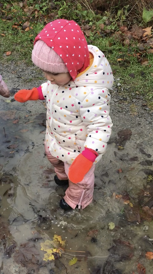 child in white jacket, red gloves and hood standing in a puddle