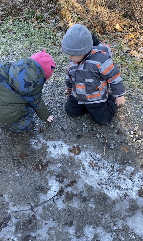 Two children looking at puddles that have turned to ice, with one of them pointing down