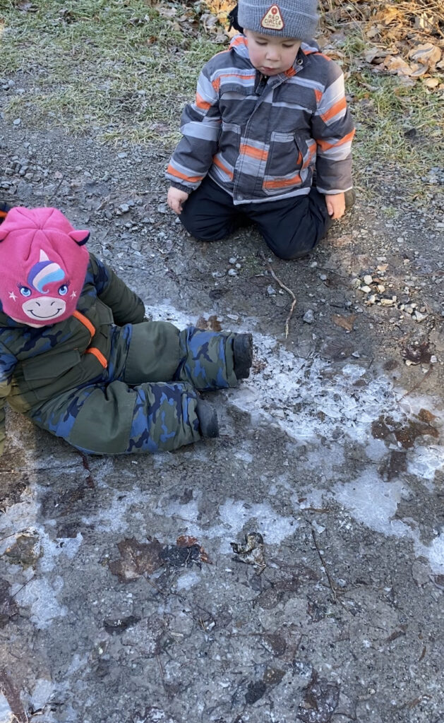 Two children sitting on the ground looking at puddles that have turned to ice