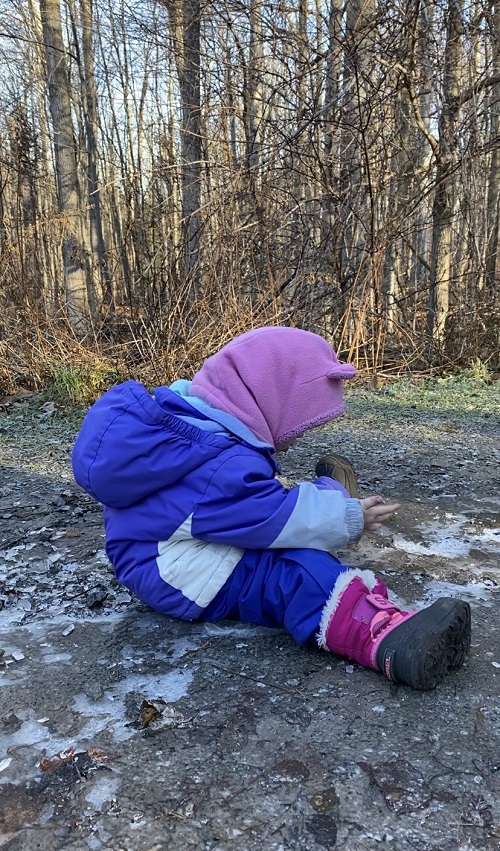 Child in a pink hat and blue jacket sitting on the icey ground looking down