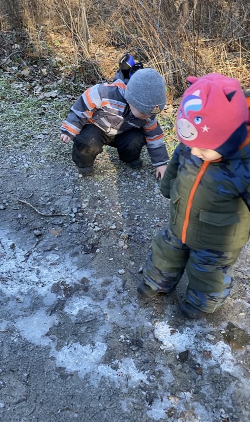 Two children, one standing and one squatting, looking at frozen puddles on the ground