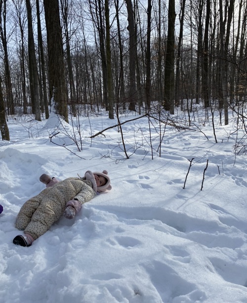 Little girl in pink hat and white patterned snowsuit laying in the snow in a forest.