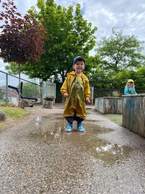Small boy outside in a yellow rain jacket playing in a puddle on a rainy day.