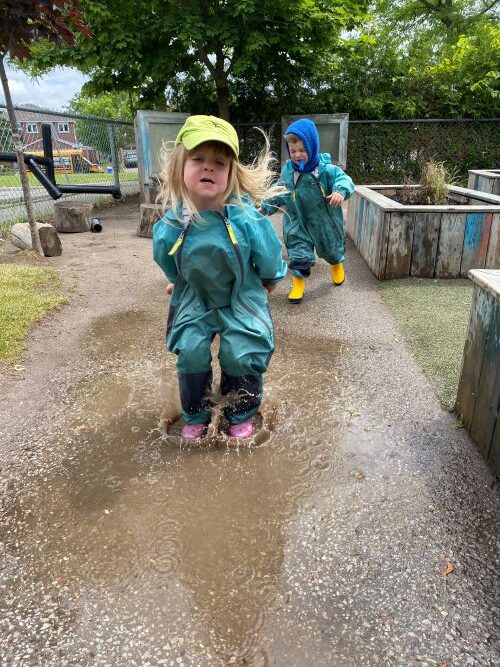 A small girl outside in a teal rain jacket and yellow hat, playing in a puddle on a rainy day.
