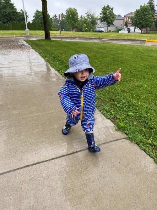 Boy in blue rain gear walking on wet sidewalk, pointing up.