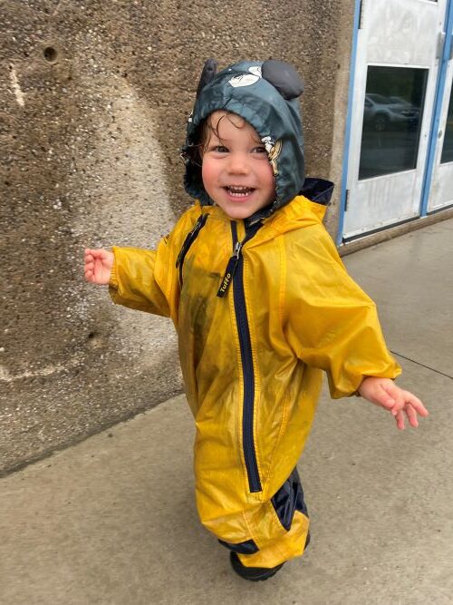 Child in yellow rain jacket and green hat smiling outside on a rainy day.
