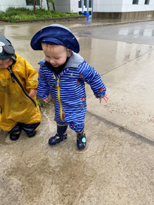 Toddler in blue rain gear standing beside a child in yellow rain jacket on the wet sidewalk.