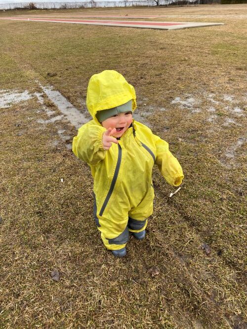 Infant in yellow rain suit standing on the wet grass.