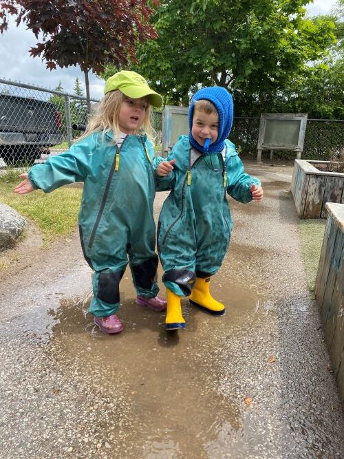 Two children in teal rain jackets playin outside on a rainy day.