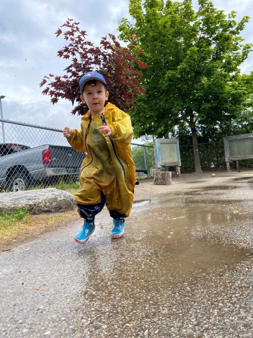 Small boy in a yellow rain jacket and blue hat playing outside on a rainy day.