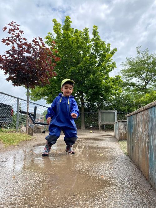 Small boy in a blue rain jacket playing outside in a puddle on a rainy day.