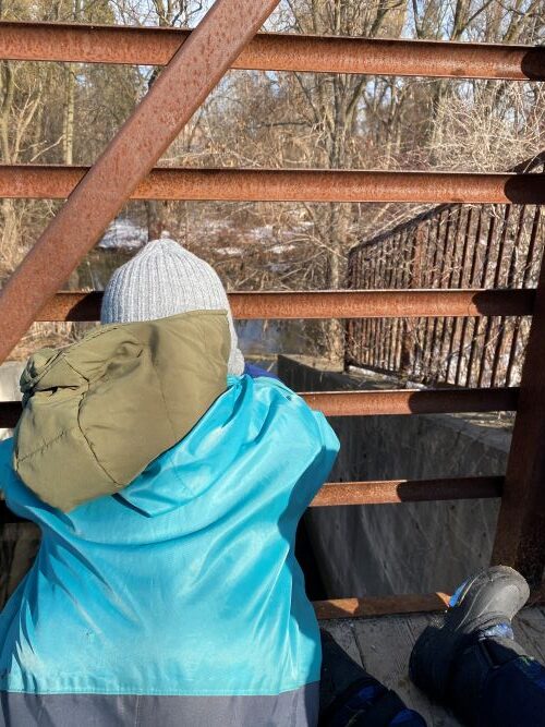 Child in teal jacket looking out from a metal bridge.