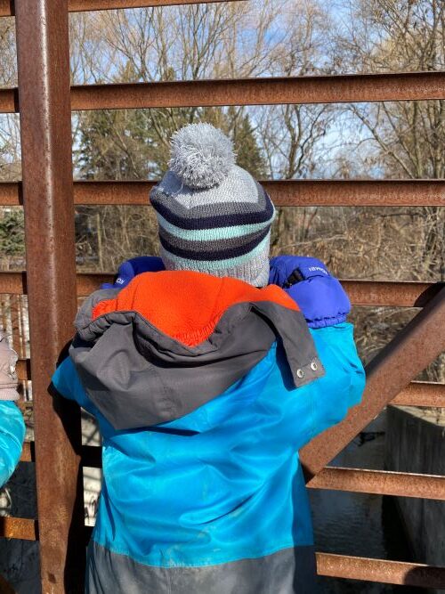 Child in blue jacket and grey hat looking out from a metal bridge.