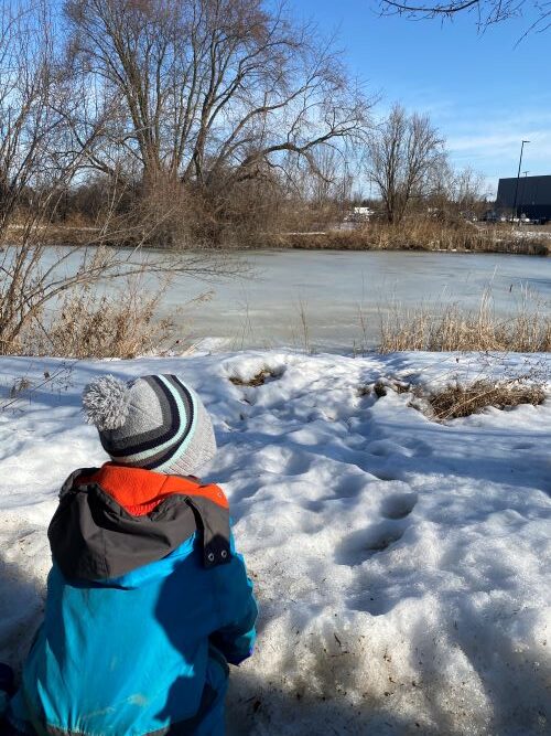 Boy in hat and jacket outside looking out at the snow and water.