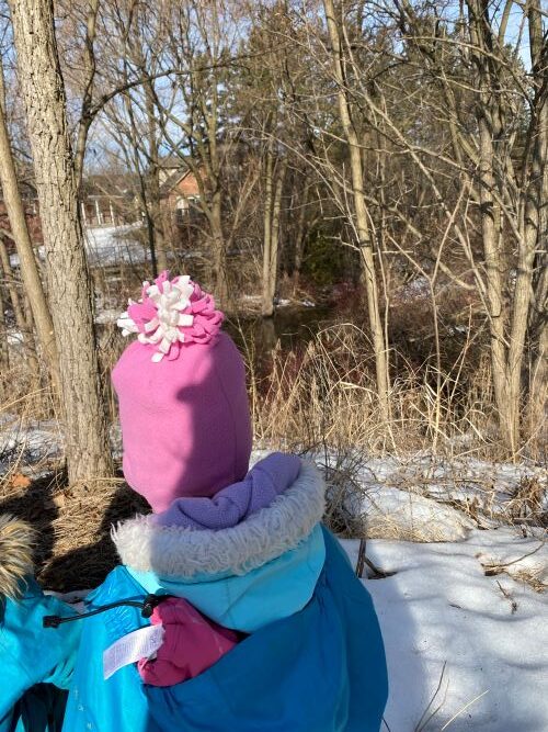 View of child from the back wearing pink hat and blue jacket walking on a path in the winter.