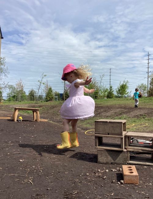 Girl in pink hat and dress, with yellow rain boots jumping off small box.